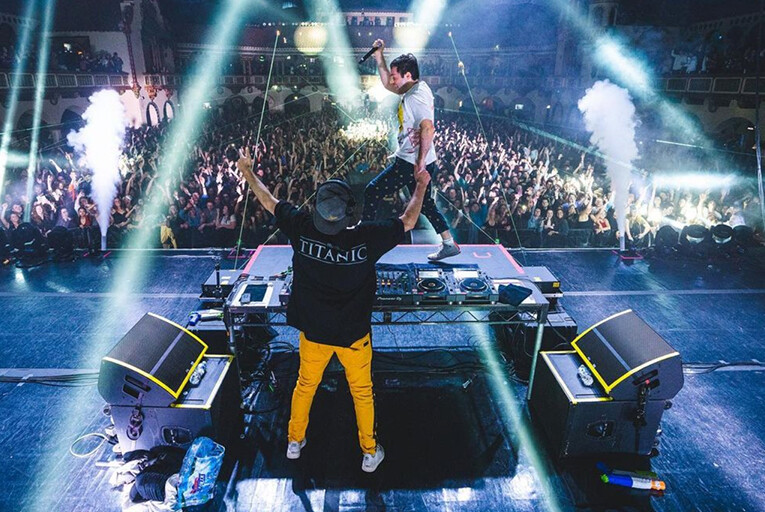 Two Friends performing in front of a large crowd. One is looking at the crowd, the other is standing on the DJ table.