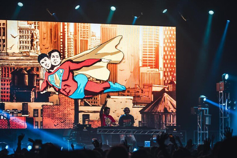 Two Friends live in concert in front of a giant backdrop depicting them as a conjoined superhero.